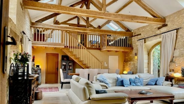 A spacious living room with a high ceiling, exposed wooden beams, a staircase leading to an upper level, and a cozy seating area with a sofa and armchairs.