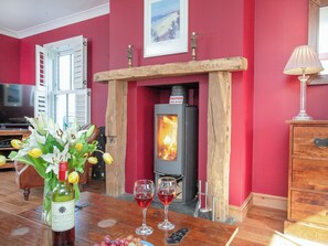 A cozy living room with a wood-burning stove, a wooden mantelpiece, a table with wine glasses and a bottle, and a television in the background.