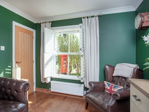 Ein Zimmer mit grüner Wand, einer Holztür, einem Fenster mit weißen Vorhängen, einem braunen Ledersofa und einem Kissen mit Union Jack sowie einem Heizkörper.