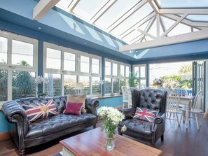 A conservatory with a glass ceiling, two leather sofas with Union Jack cushions, a wooden table, and a view of the garden outside.