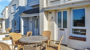 Un patio con mobiliario de mimbre y una mesa redonda, junto a un edificio de ladrillo blanco con una puerta y ventanas.