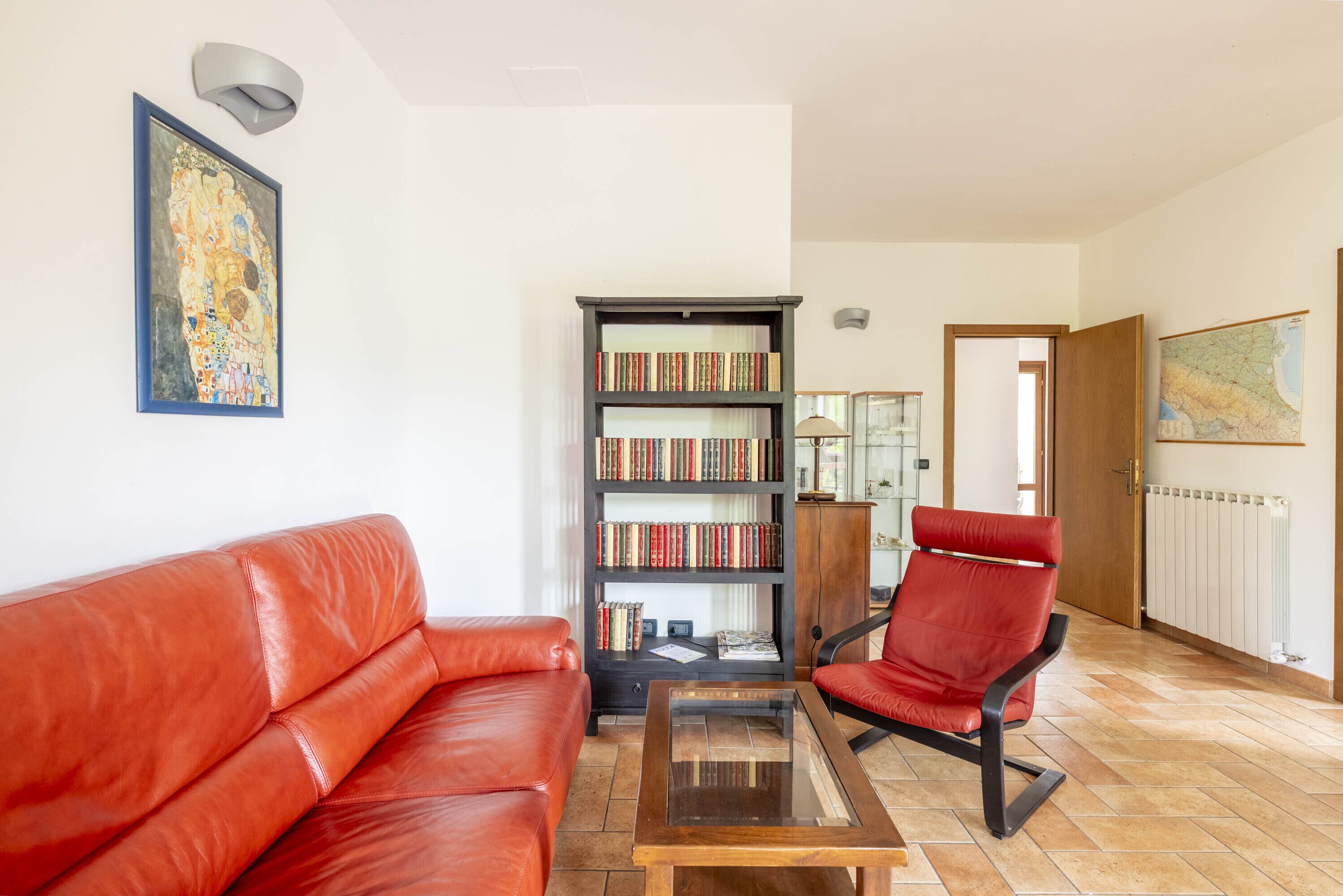 Een woonkamer met een rode leren bank, een houten bijzettafeltje, een rode fauteuil, een boekenkast en een lijst met een schilderij aan de muur.