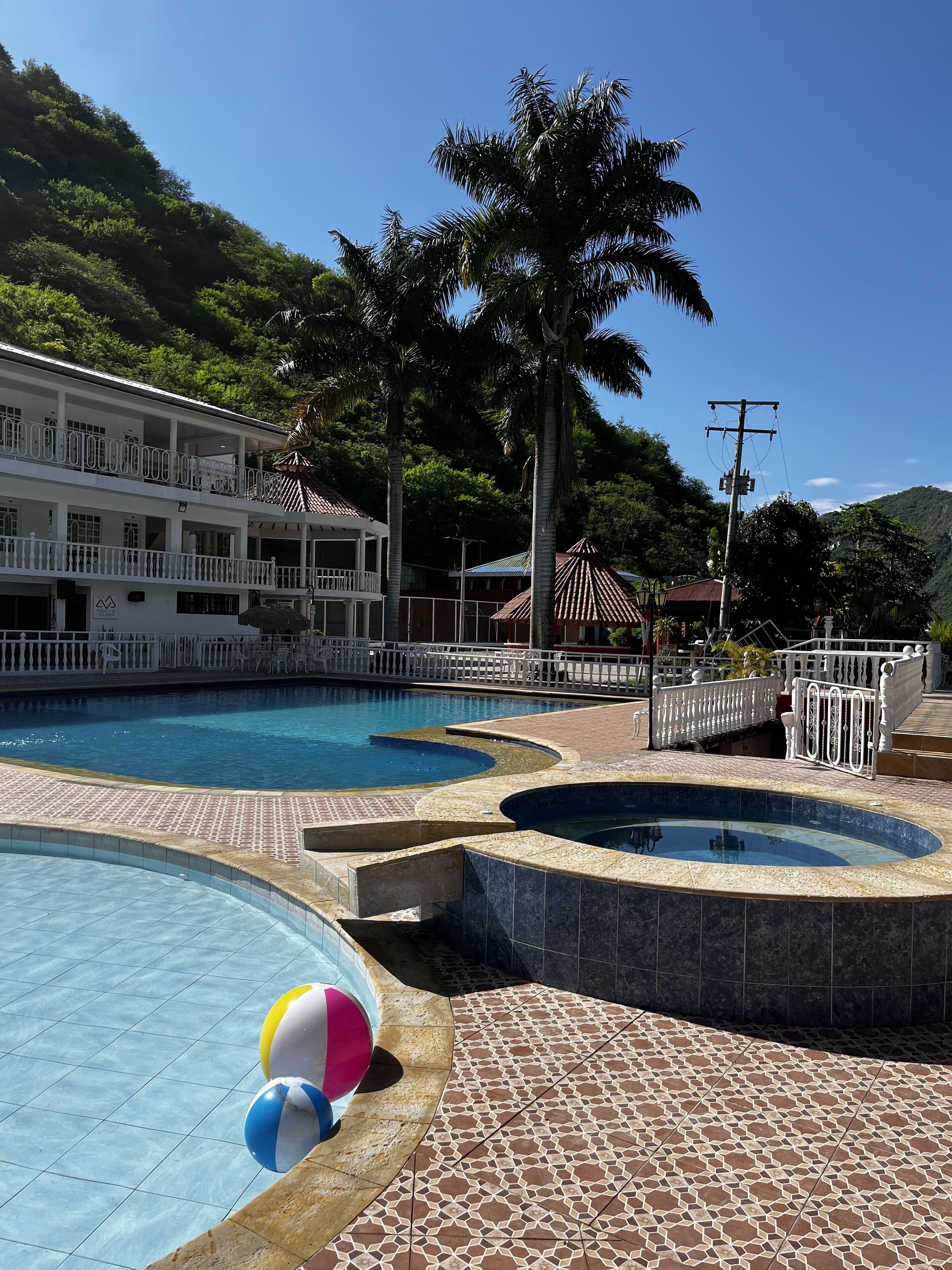 Uma área de piscina com diversas fontes, um edifício branco com varandas e vegetação ao redor.