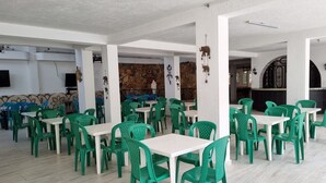 A spacious indoor dining area with green plastic chairs and white tables, white walls, and a stone wall decoration.