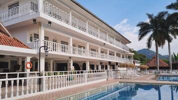 Un hotel de varios pisos con piscina, balcones y montañas al fondo.