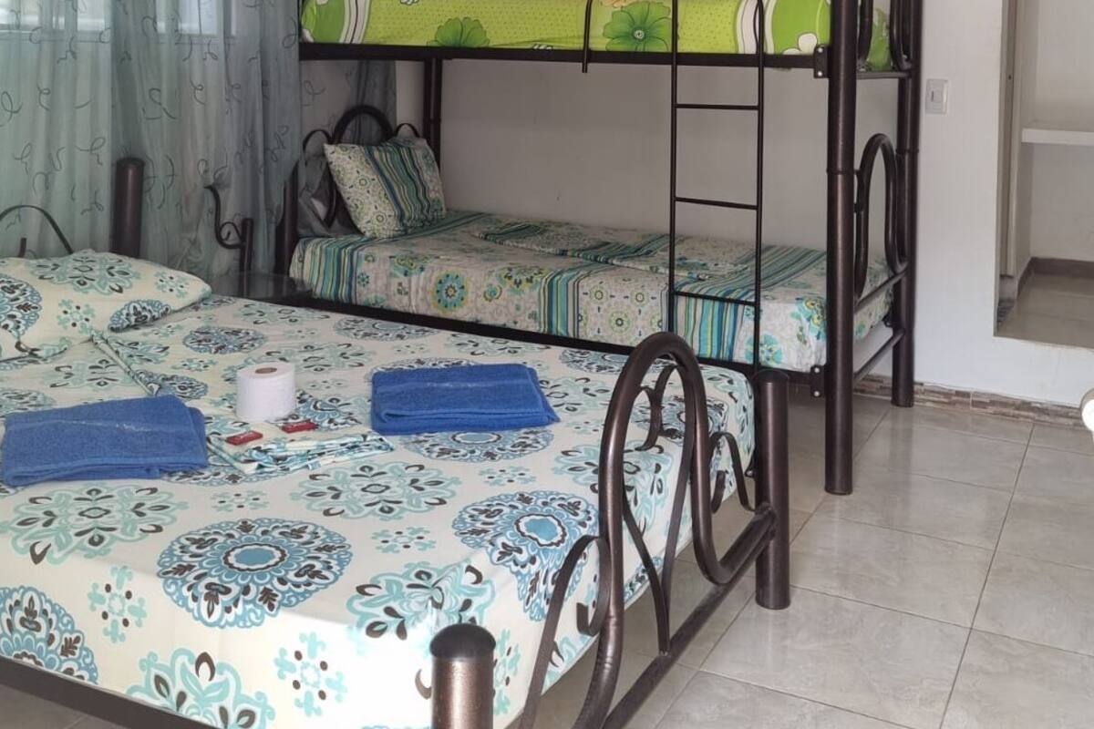 A dormitory room with bunk beds, a window with curtains, and a bed with a patterned bedspread.