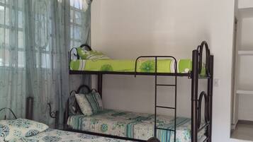 A dormitory room with bunk beds, a window with curtains, and a bed with a patterned bedspread.