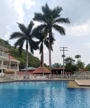 A swimming pool with palm trees, a covered seating area, and a building with a red roof. - Hotel Mirador de la Montaña (Anapoima)