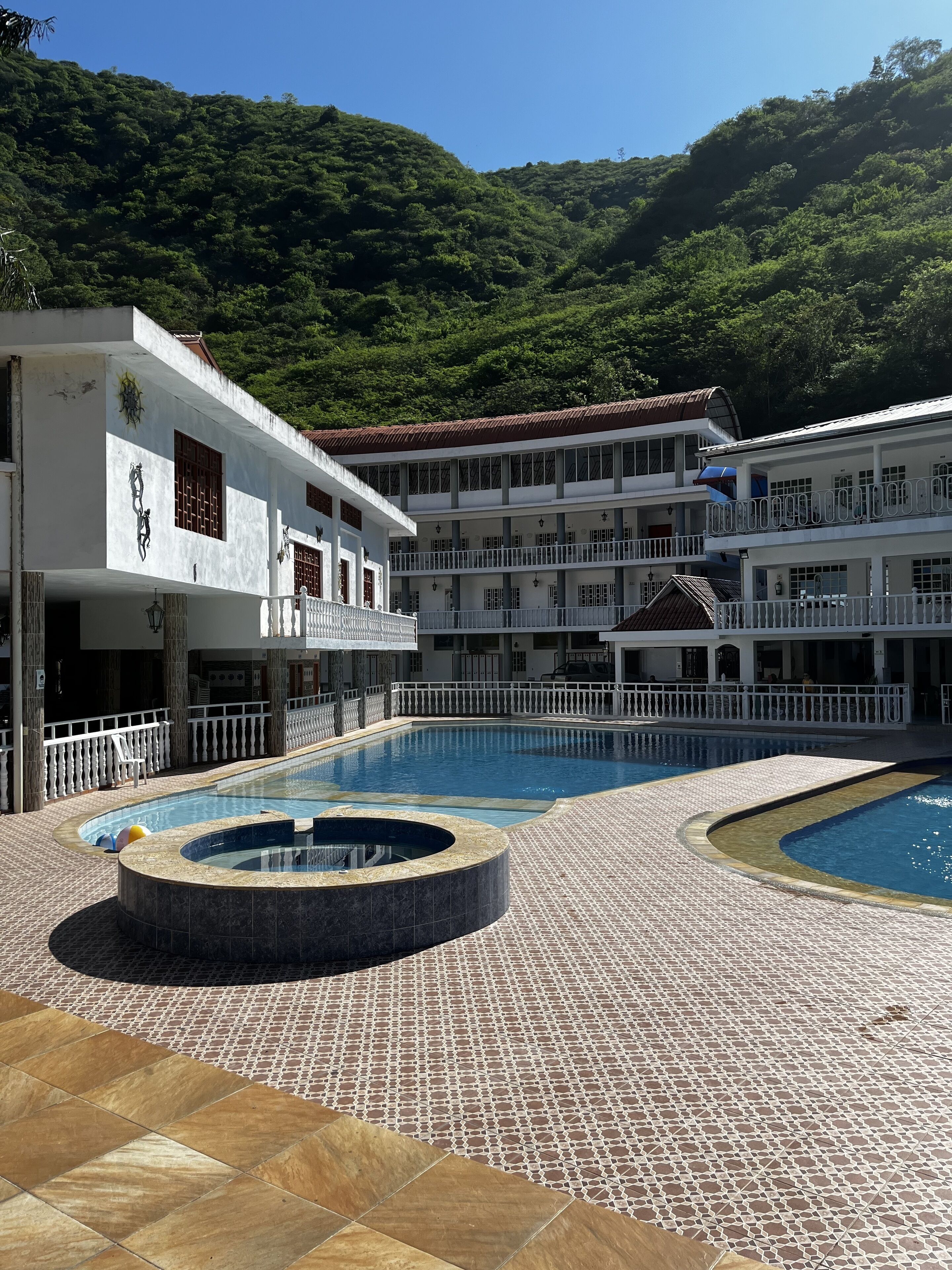 Um hotel com uma grande piscina ao ar livre, várias varandas e montanhas ao fundo.