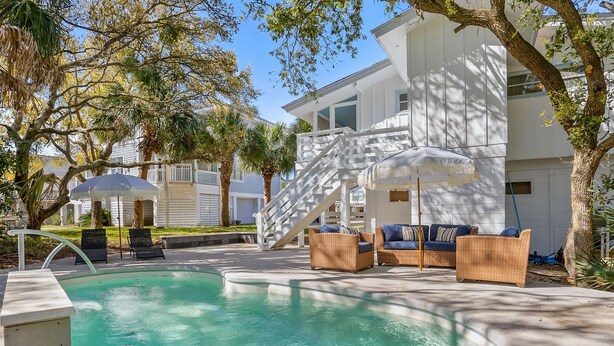 A swimming pool with lounge chairs and umbrellas, surrounded by a white building with a staircase and a covered area. - Private Pool-Fripp Island-Steps From The Beach (Fripp Island)