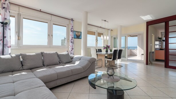A modern living room with a sectional sofa, a glass coffee table, and a dining area with a view of the sea.