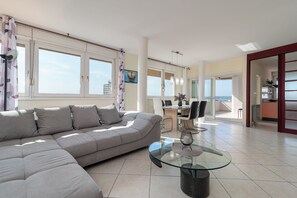 A modern living room with a sectional sofa, a glass coffee table, and a dining area with a view of the sea.