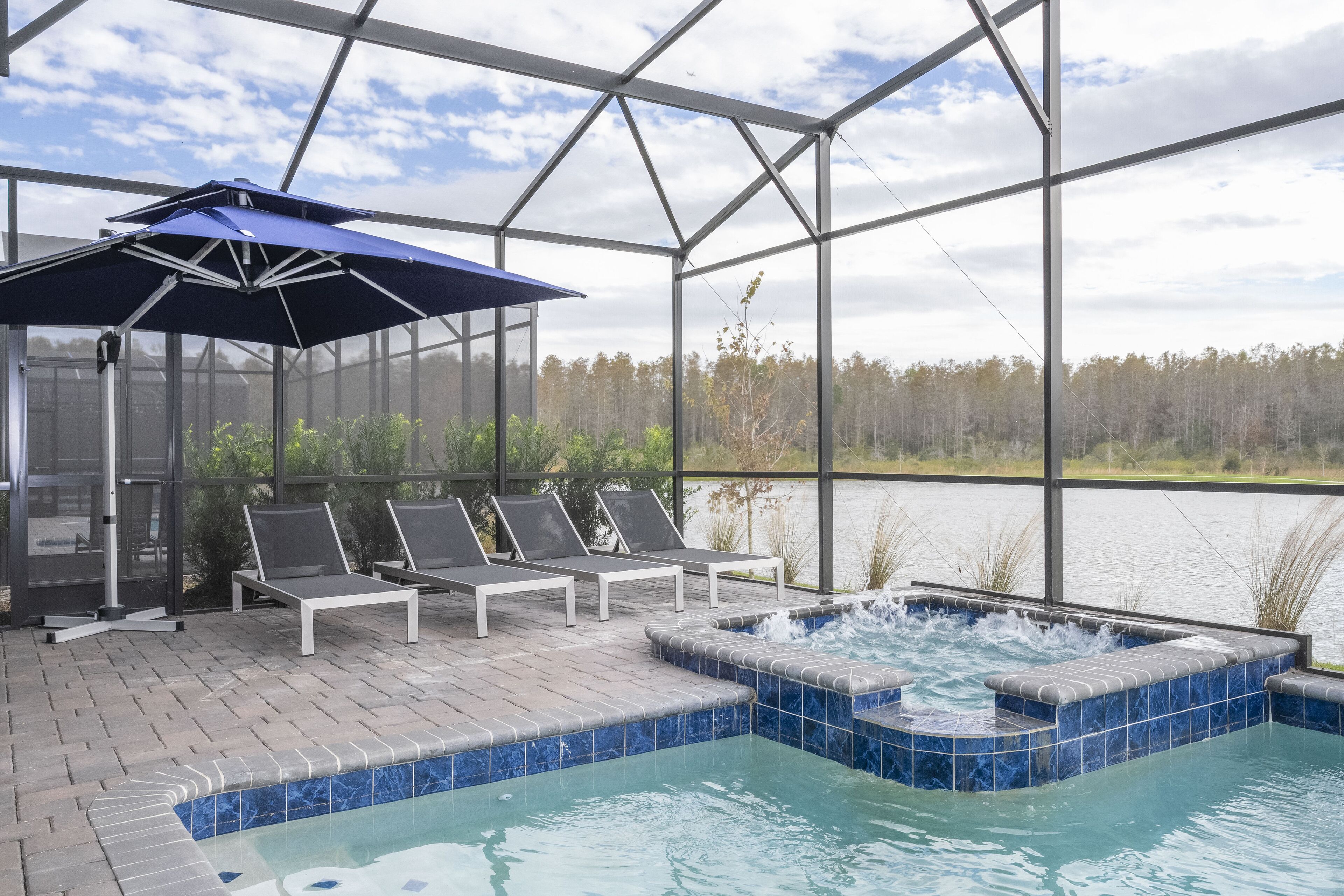 A covered outdoor pool area with lounge chairs and a hot tub, overlooking a body of water and forest.