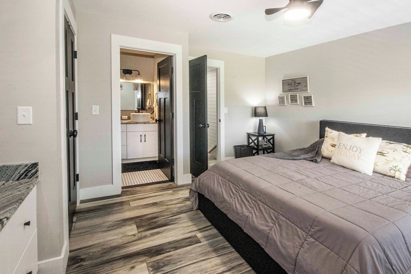 Un dormitorio con cama, mesita de noche, lámpara, puerta que da al baño y ventilador de techo.