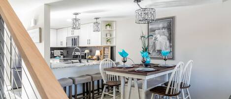 Una cocina moderna con armarios blancos, un comedor con una mesa de madera y sillas blancas, y una escalera con barandilla de madera.