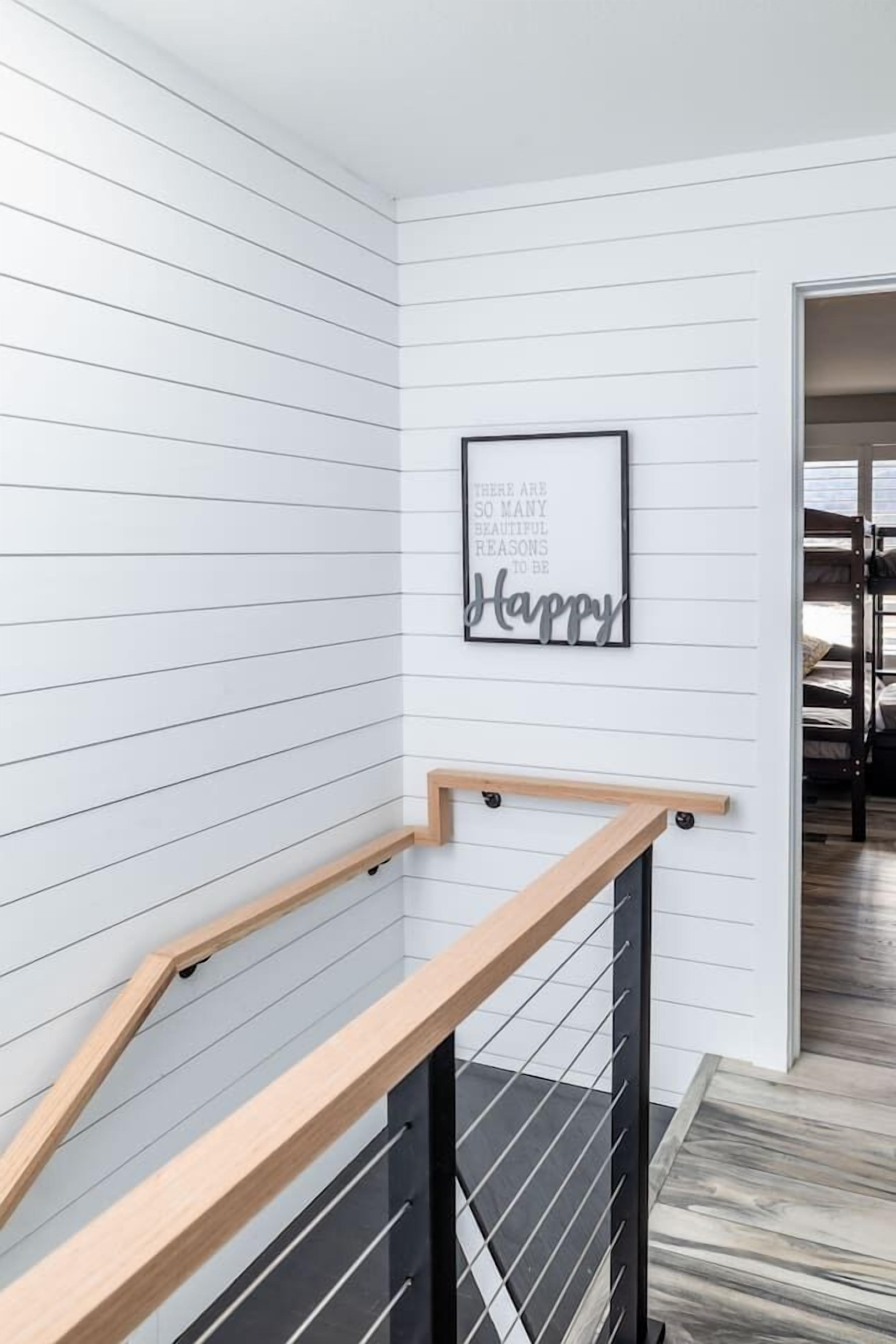 Un escalier avec des rampes en bois et une balustrade en métal noir, des murs recouverts de panneaux blancs et une citation encadrée accrochée au mur.