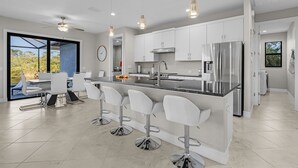 A modern kitchen with white cabinets, stainless steel appliances, and a bar area with white stools. - Brandnew (2024) Villa with huge pool on the water (Port Charlotte)