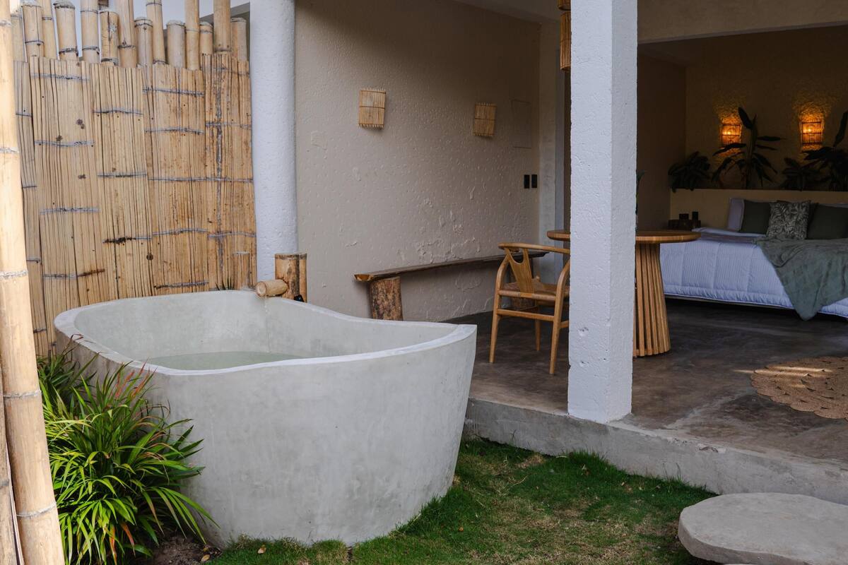 Un área de spa con una bañera grande, una silla de madera, una mesita y una cama con vista a las plantas.