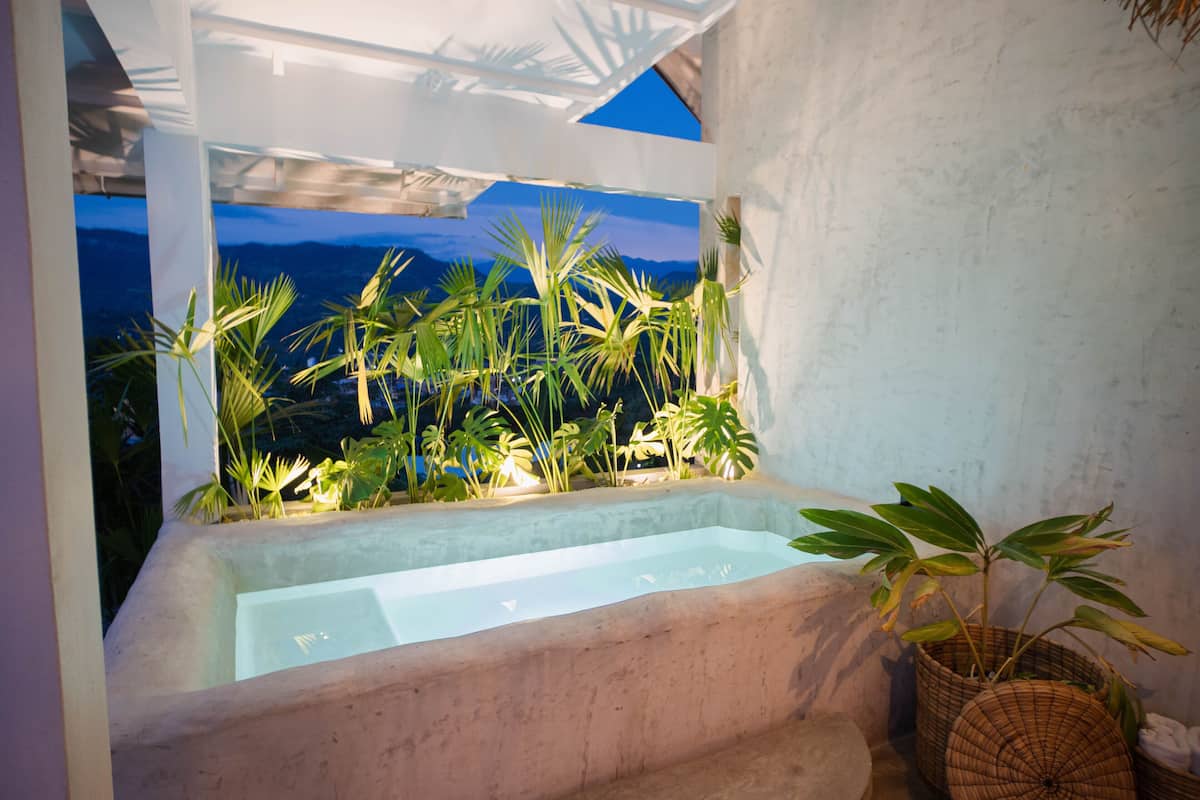 Un área de spa con un jacuzzi, rodeado de plantas tropicales y vistas a las montañas al anochecer.