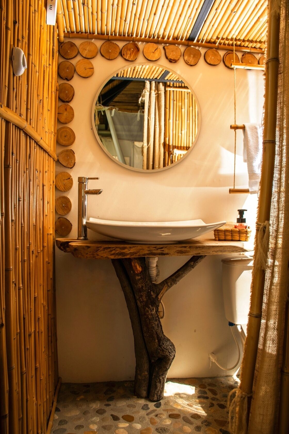 Un bagno con parete in bambù, uno specchio rotondo, un lavabo fissato a un ceppo d'albero e un water.