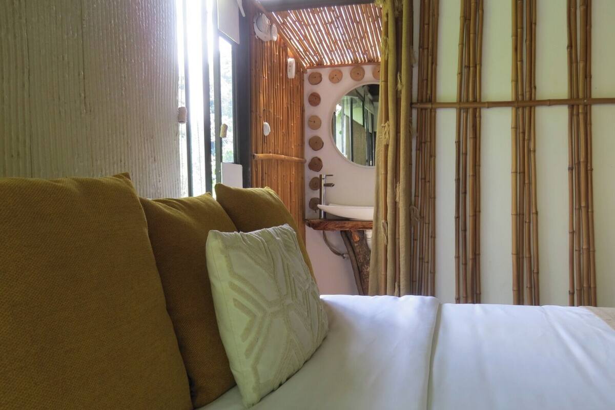 Una cama con almohadas, un baño con lavamanos y espejo, y paneles de pared de bambú.