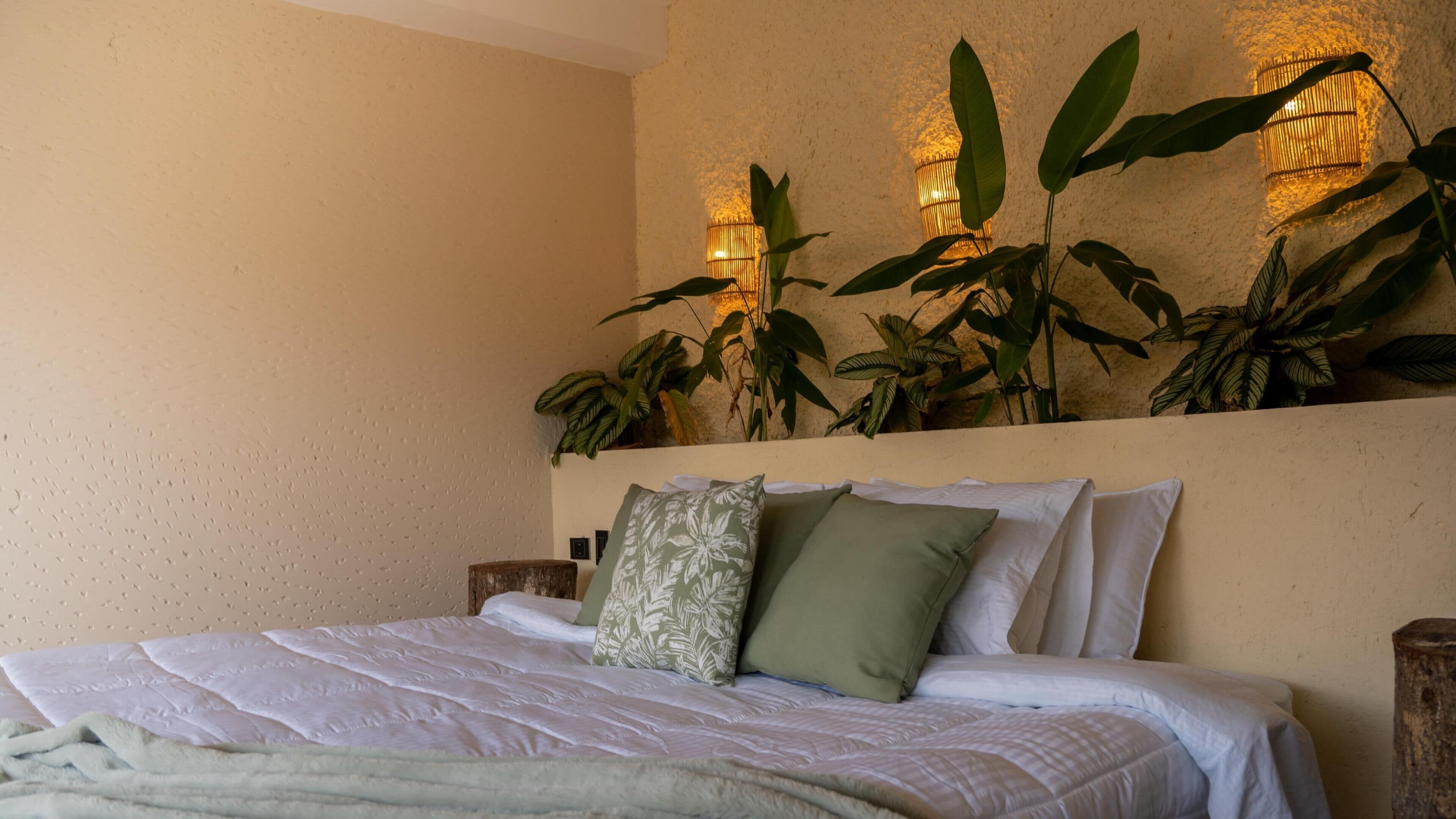 A bed with white bedding, a green pillow, and a patterned pillow, with large green plants and wall-mounted lights in the background.