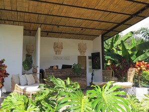 A covered outdoor seating area with wicker furniture, a reception desk, and tropical plants. - ECO HOTEL VERANO AZUL (Villeta)