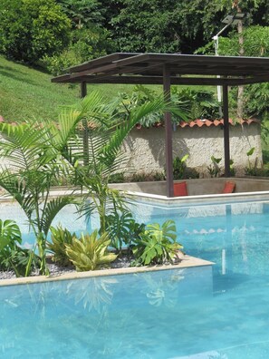 A swimming pool with a pergola, surrounded by tropical plants and a stone wall. - ECO HOTEL VERANO AZUL (Villeta)