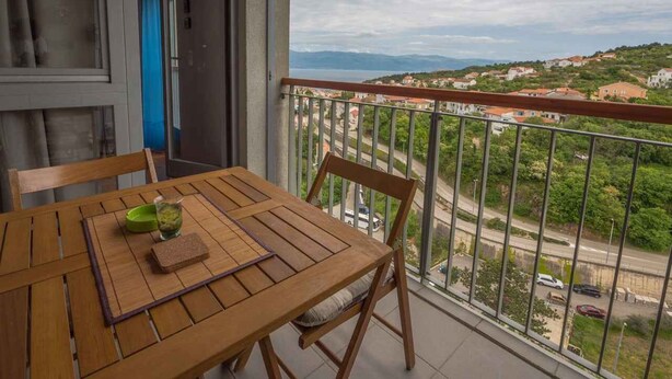 A balcony with a wooden table and chairs, a view of a road and buildings, and a door leading to an interior space. - Holiday apartment with partial sea view (Vrbnik)