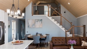 Un vaste espace de vie aménagé dans un style loft, comprenant une table à manger, des sièges, un escalier et une cuisine équipée d’un réfrigérateur.