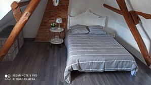 Un dormitorio con techo inclinado, una cama con ropa de cama a rayas, una pequeña mesa redonda con una lámpara y una pared de ladrillo.