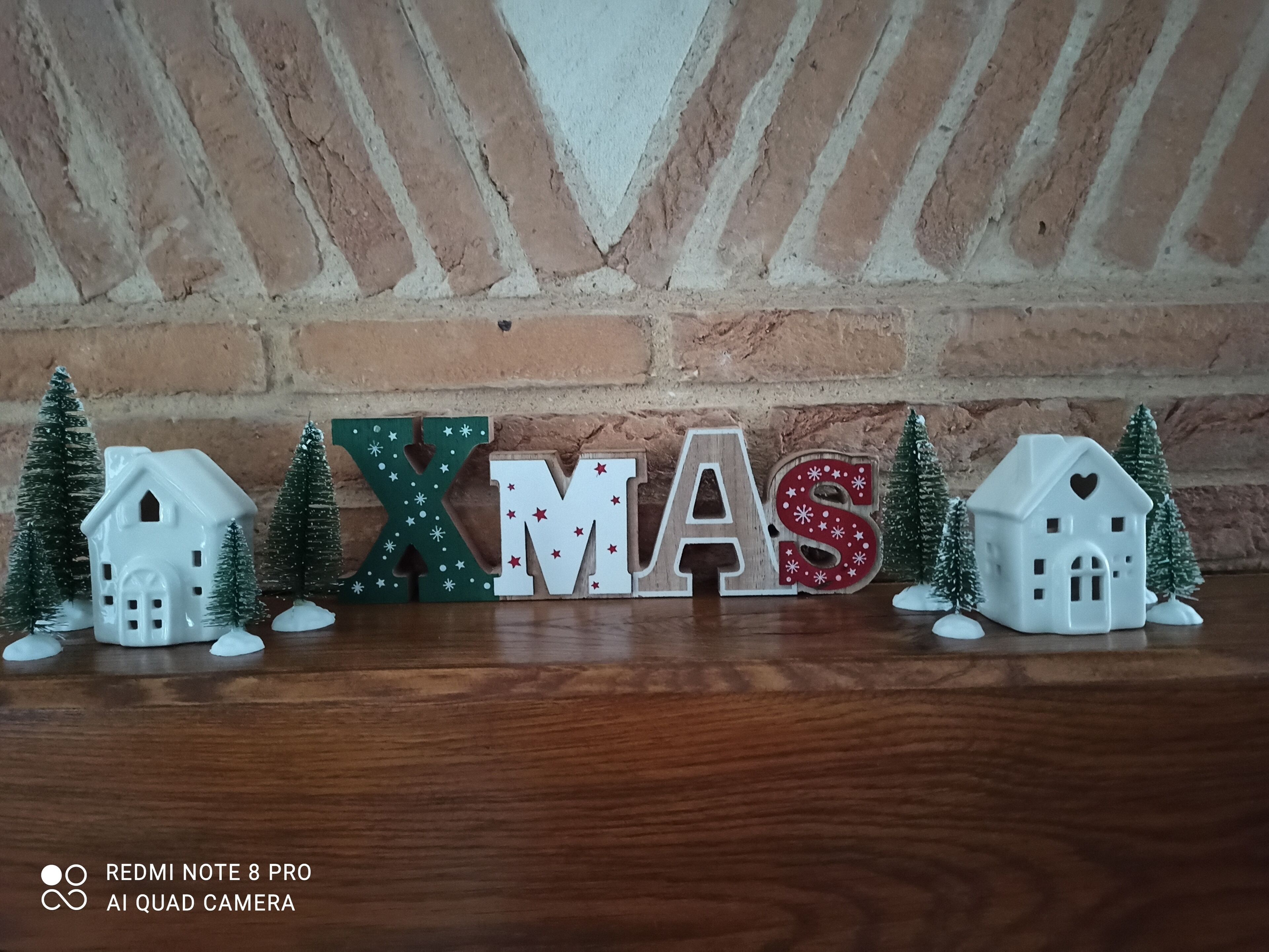 A wooden table with a brick wall background, decorated with Christmas-themed letter cutouts spelling "XMAS" and small house figurines.