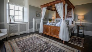 A four-poster bed with white linens, a wooden headboard, and a canopy. There is a large window with white shutters, a small table with a lamp, and a patterned rug on the floor. - House of the Northern Gate (Thurso)