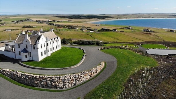 A white building with a curved driveway and a stone wall, surrounded by green fields and a body of water. - House of the Northern Gate (Thurso)