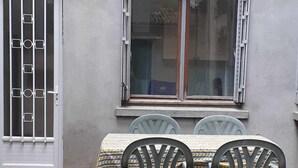 Een terras met een tafel en vier stoelen, een raam met open luiken en een metalen hek.