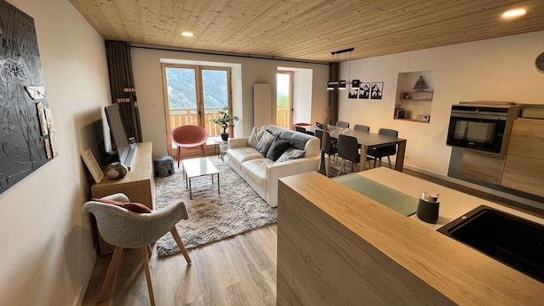 A modern living space with a wooden ceiling, a dining area, a kitchen with a built-in oven, and large windows offering a mountain view.