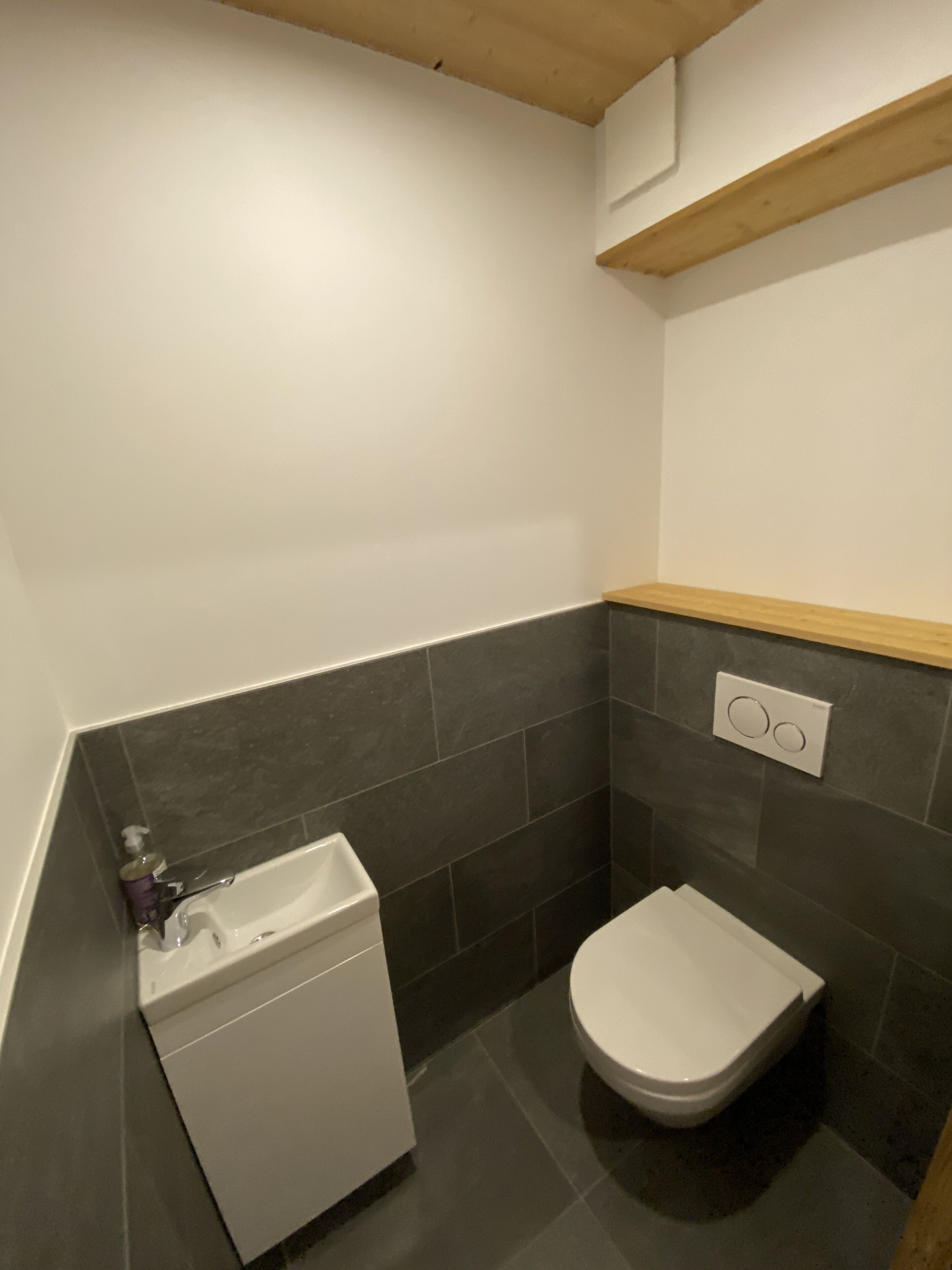 Une petite salle de bain avec des toilettes blanches, un lavabo blanc et une étagère en bois au-dessus des toilettes.