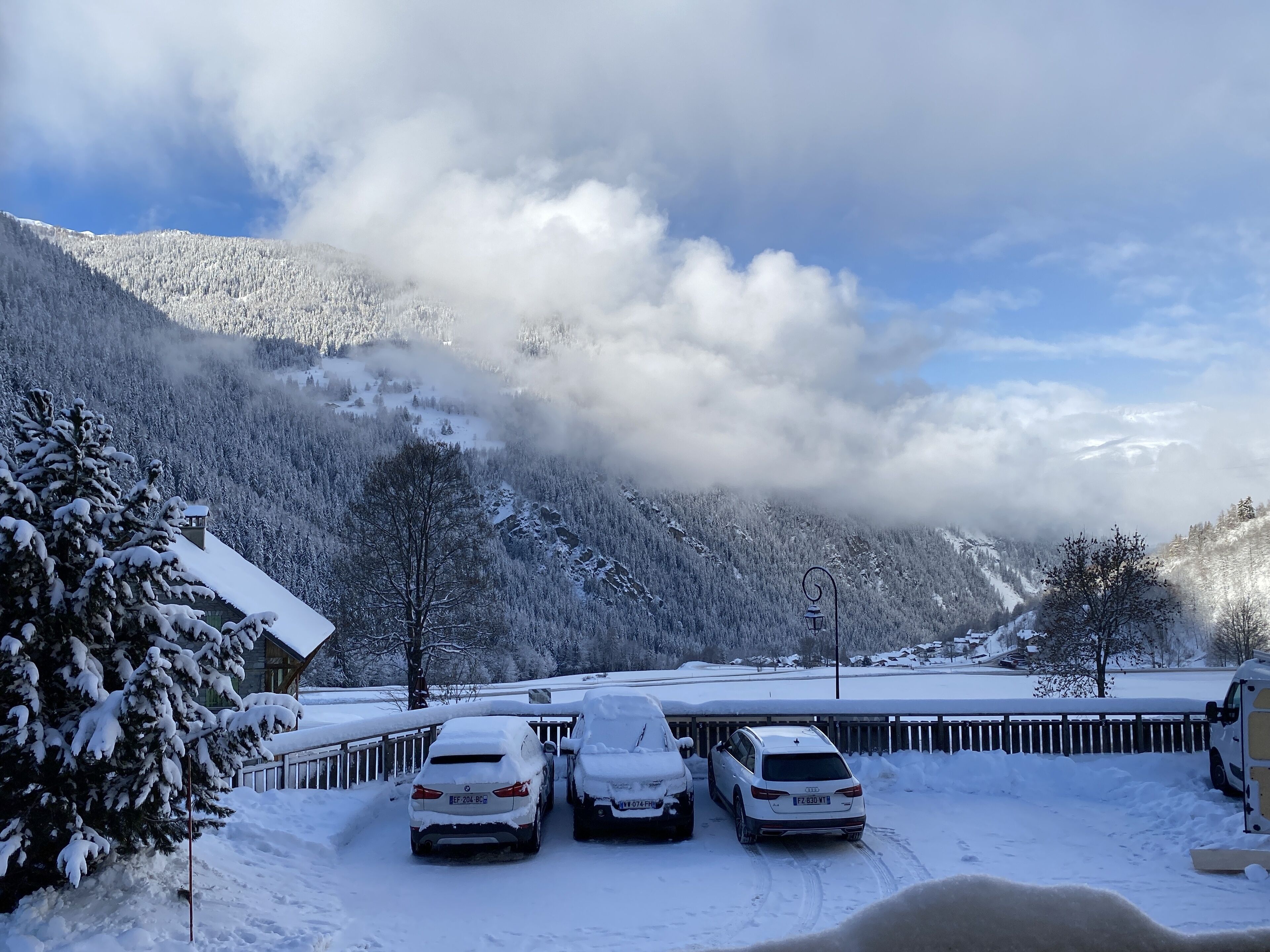 Três carros estacionados em uma área nevada, com montanhas ao fundo e uma cerca de madeira.
