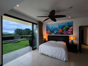 Une chambre avec un grand lit, un ventilateur de plafond, un tableau et une vue sur un jardin.