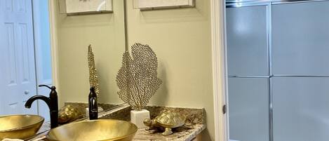 A bathroom with a vanity, a large gold bowl sink, a mirror, and a shower area.