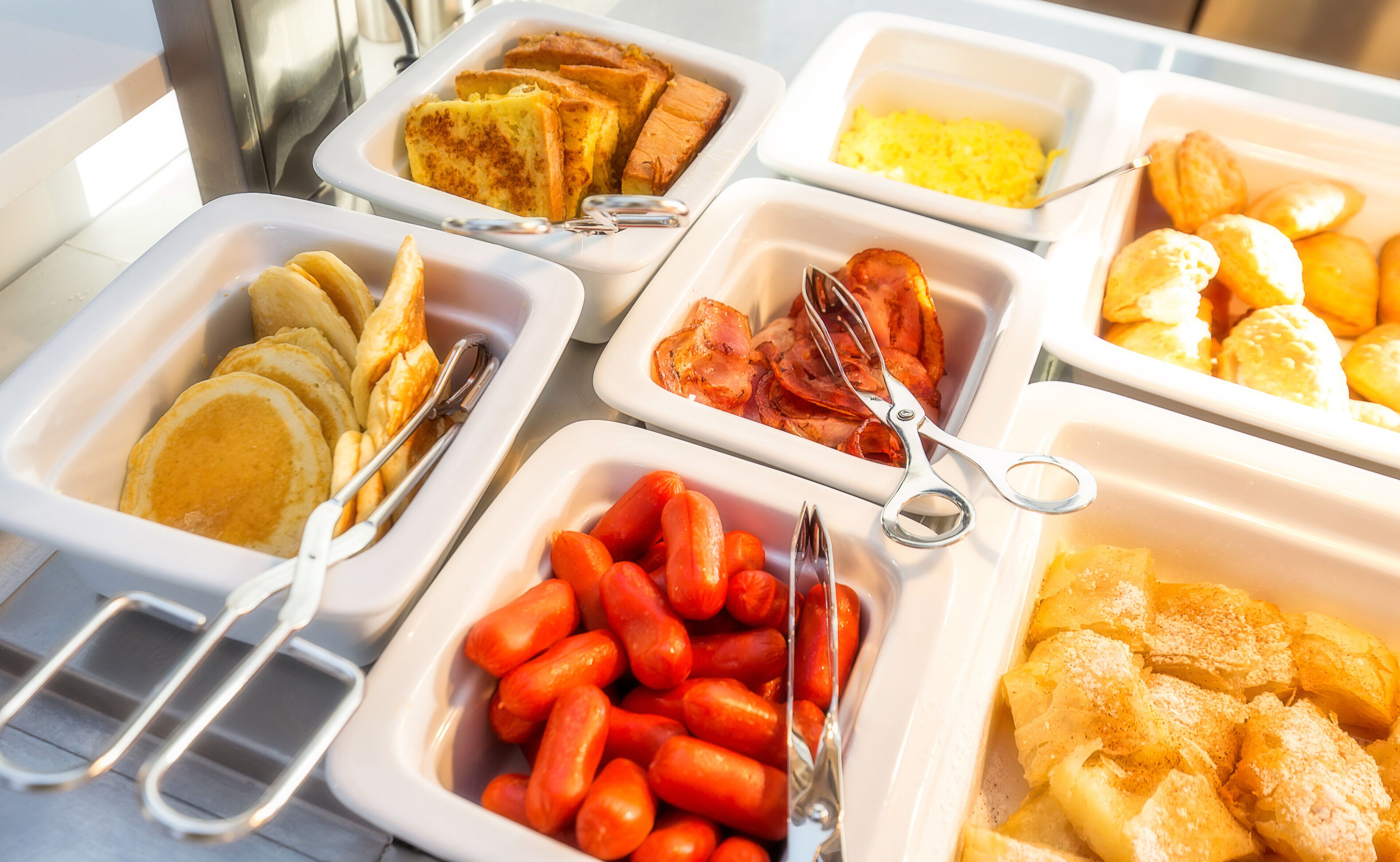 A buffet with various food items including pancakes, bacon, sausages, and pastries.