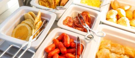 Un bufé con diversas opciones de comida, incluyendo panqueques, tocino, salchichas y pastelería.