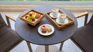 Une table ronde avec un plateau de pâtisseries, une assiette contenant une pâtisserie et une boisson, et une vue sur la verdure par une fenêtre.