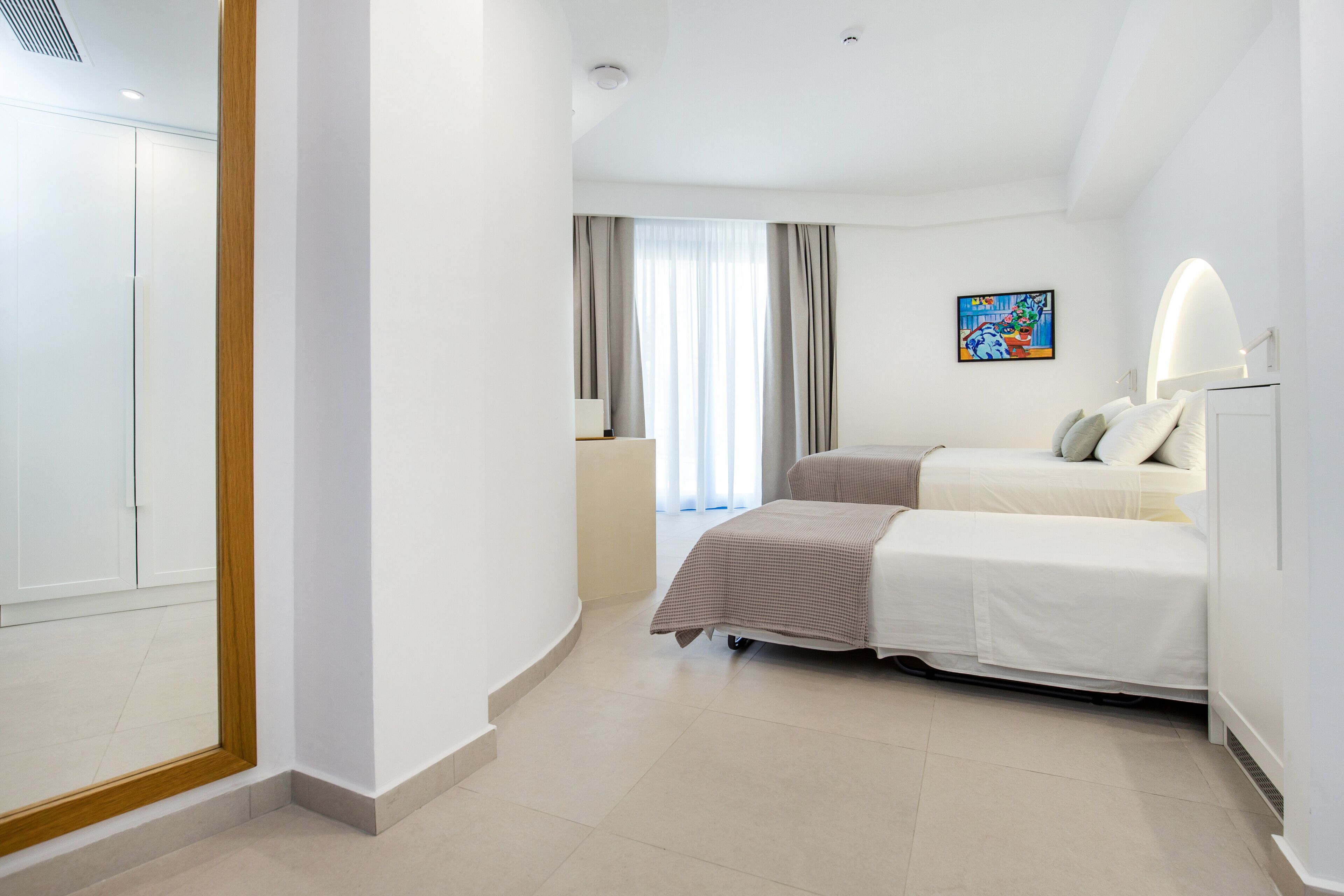 A modern hotel room with two beds, a large window with curtains, a painting on the wall, and a wooden-framed mirror.