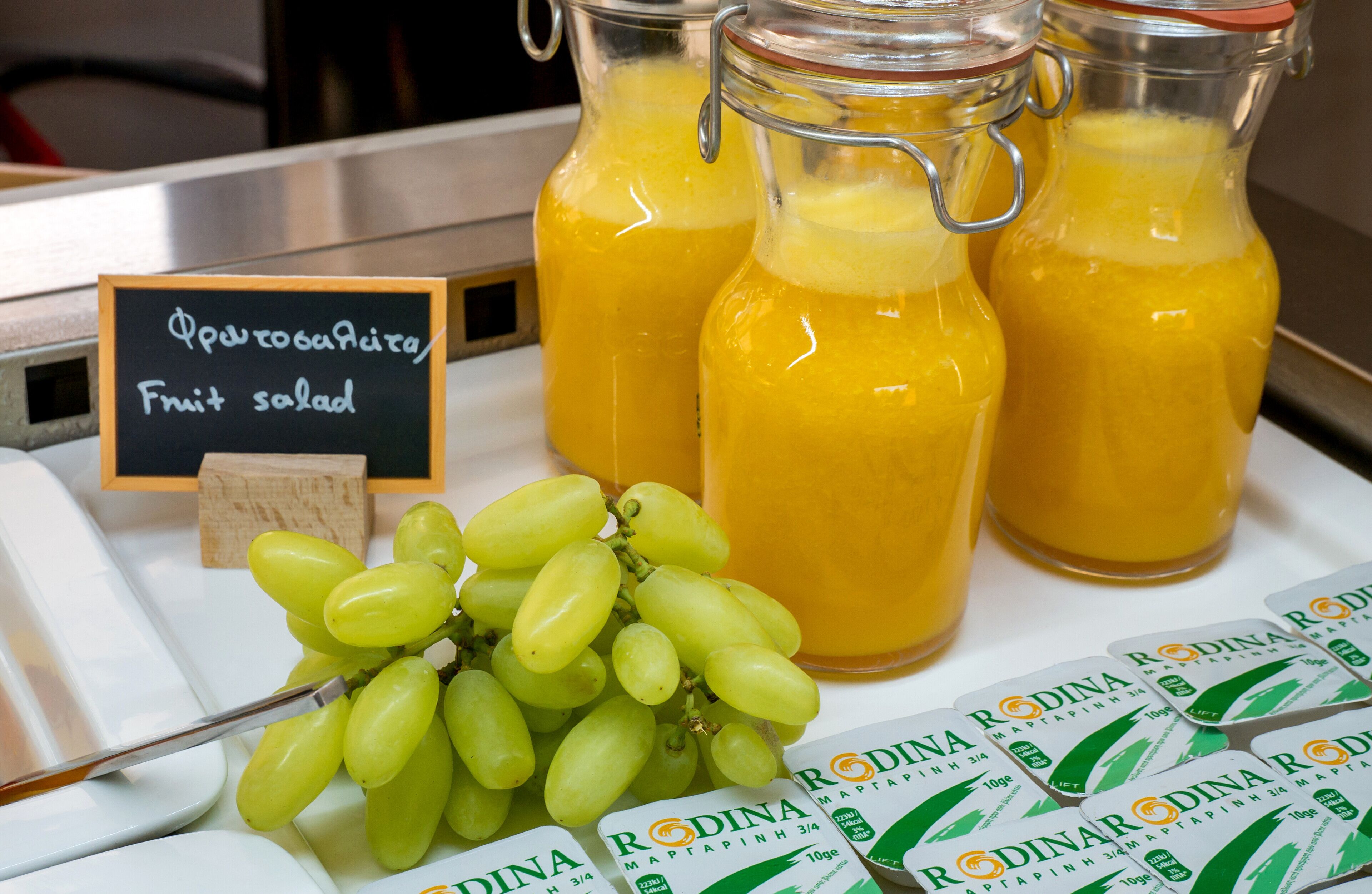 Un letrero con una ensalada de frutas, tres botellas de vidrio con jugo de naranja y un racimo de uvas verdes.