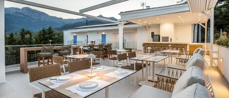 Un área de comedor en la azotea con mesas, sillas, un bar y vistas a montañas.