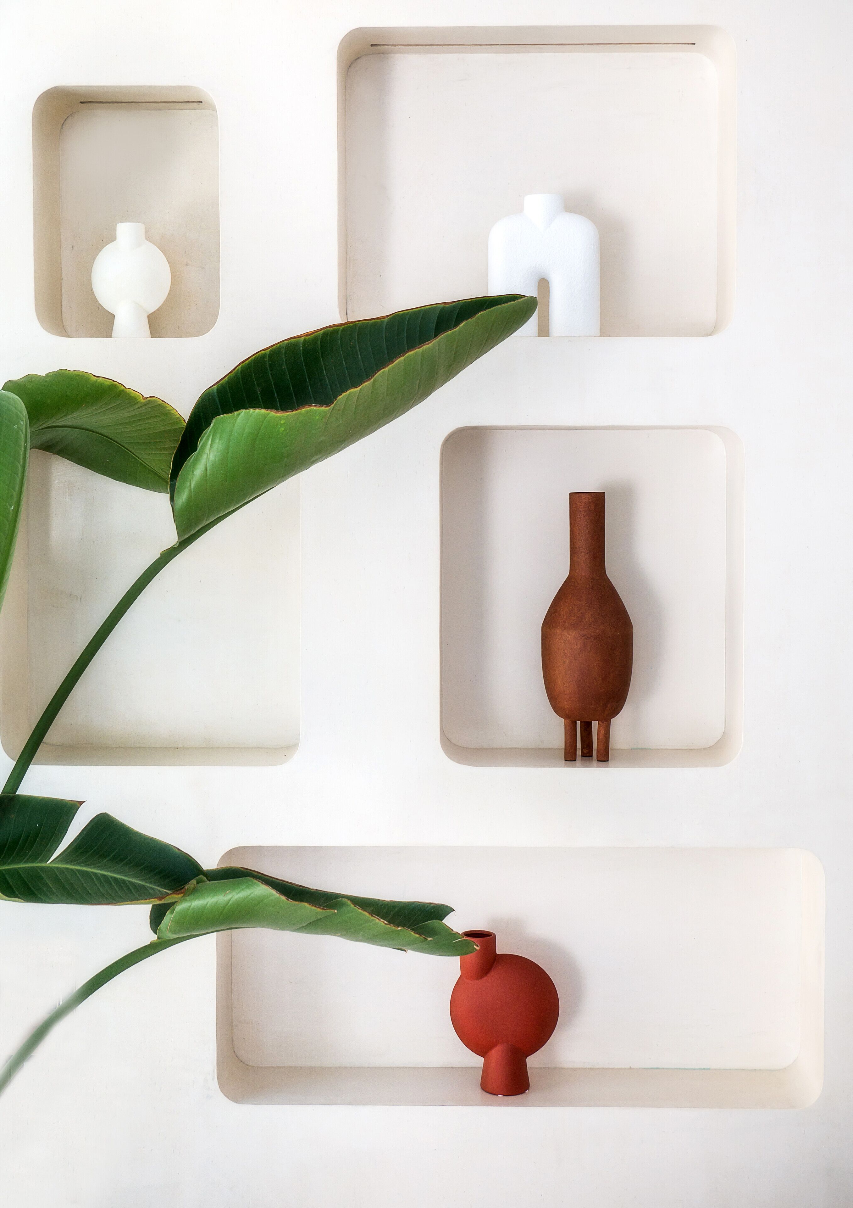 Eine schlichte Wand mit drei rechteckigen Ablagen, auf denen jeweils ein anderes Objekt steht: eine weiße Vase, eine Holzvase und eine rote Vase.