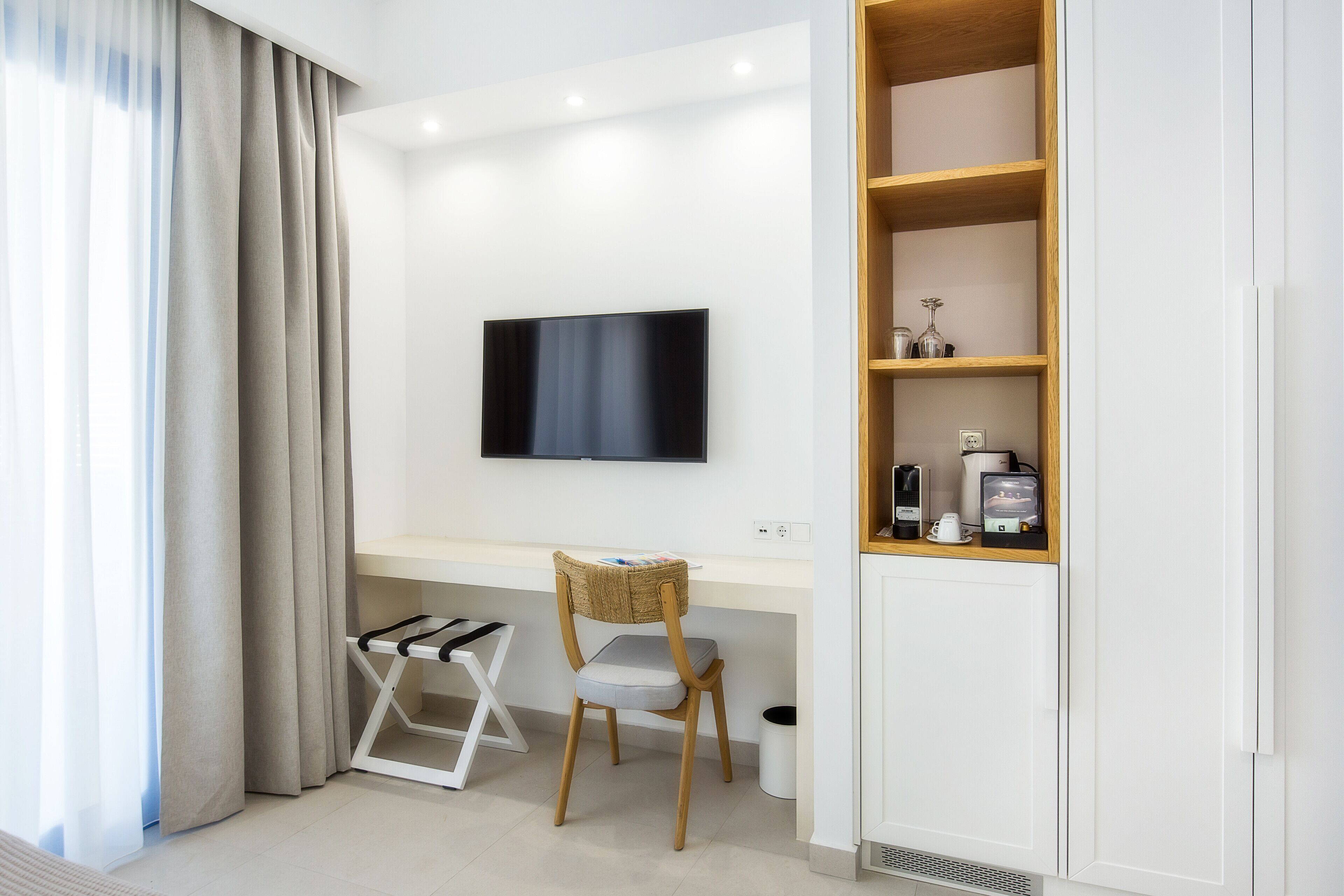 A modern room with a flat-screen TV mounted on the wall, a wooden chair, a small white desk, and a built-in wooden shelf with various items.