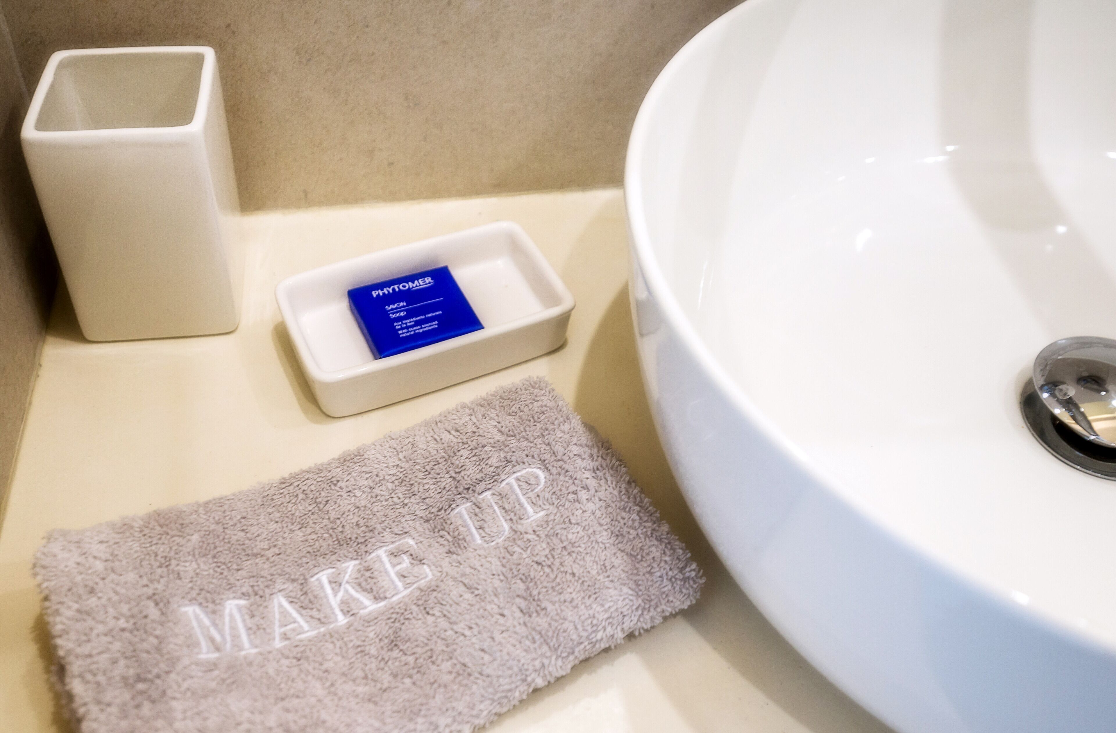Un lavamanos de baño con un lavabo blanco, un pequeño plato rectangular para jabón con etiqueta azul y una toalla con el texto 'MAKE UP'.