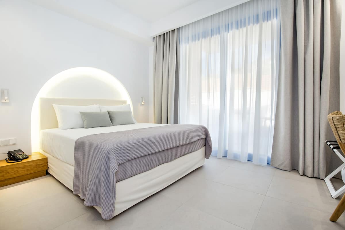 Ein modernes Hotelzimmer mit einem großen Bett, einem Nachttischtelefon und Blick ins Freie durch transparente Vorhänge.